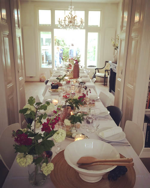 een lange tafel gedekt met wit serviesgoed, veldbloemen en lichtjes bij iemand thuis