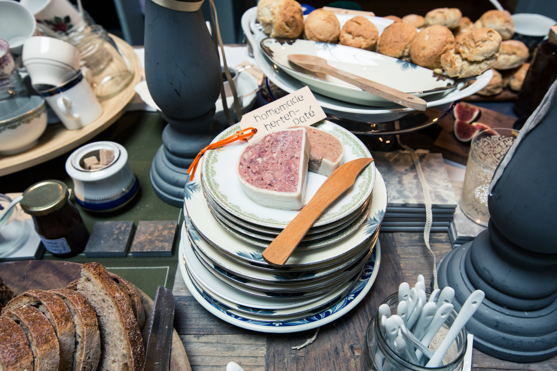 een sfeerfoto van een stoer gedekte tafel met huisgemaakte wildpate, ambachtelijk brood, houten planken, serviesgoed, scones en huisgemaakte jam